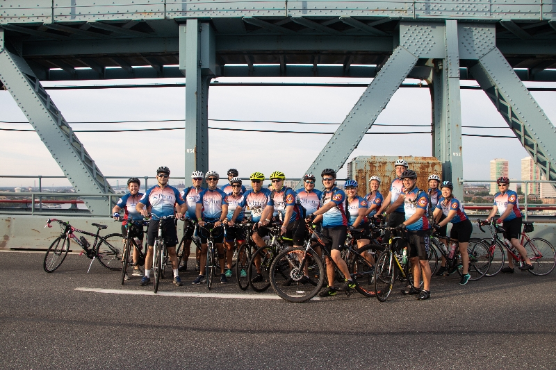 Group photo of bike riders in riding gear, with their bikes and helmets, on the Ben Franklin Bridge