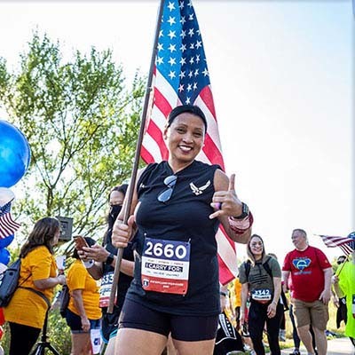 A Carry Forward female participant gives an approving hand gesture as she walks by the camera carrying the American flag during a Carry Forward 5k event.