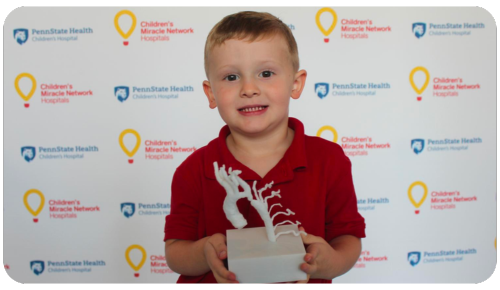 Miracle Child Henry holds a model of his 3D-printed heart in front of a Children's Miracle Network at Penn State Health Children's Hospital banner