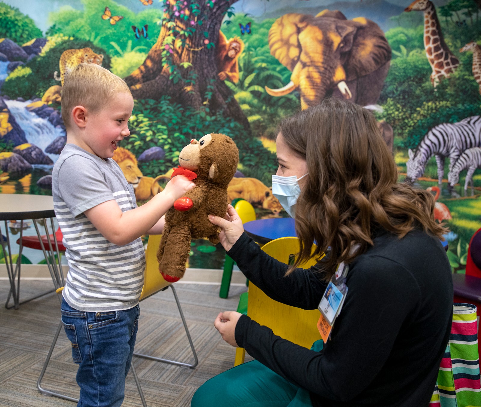 Penn State Health Child Life specialist Erin Palm hands a stuffed monkey to pediatric patient Logan in the radiology waiting room, which has a colorful safari mural on the wall